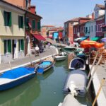 Gondole colorate attraccate a un canale di una località veneta, con caffè storici sullo sfondo.