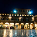 Piazza affollata nel paese marchigiano, con artigiani al lavoro e turisti che scattano foto.